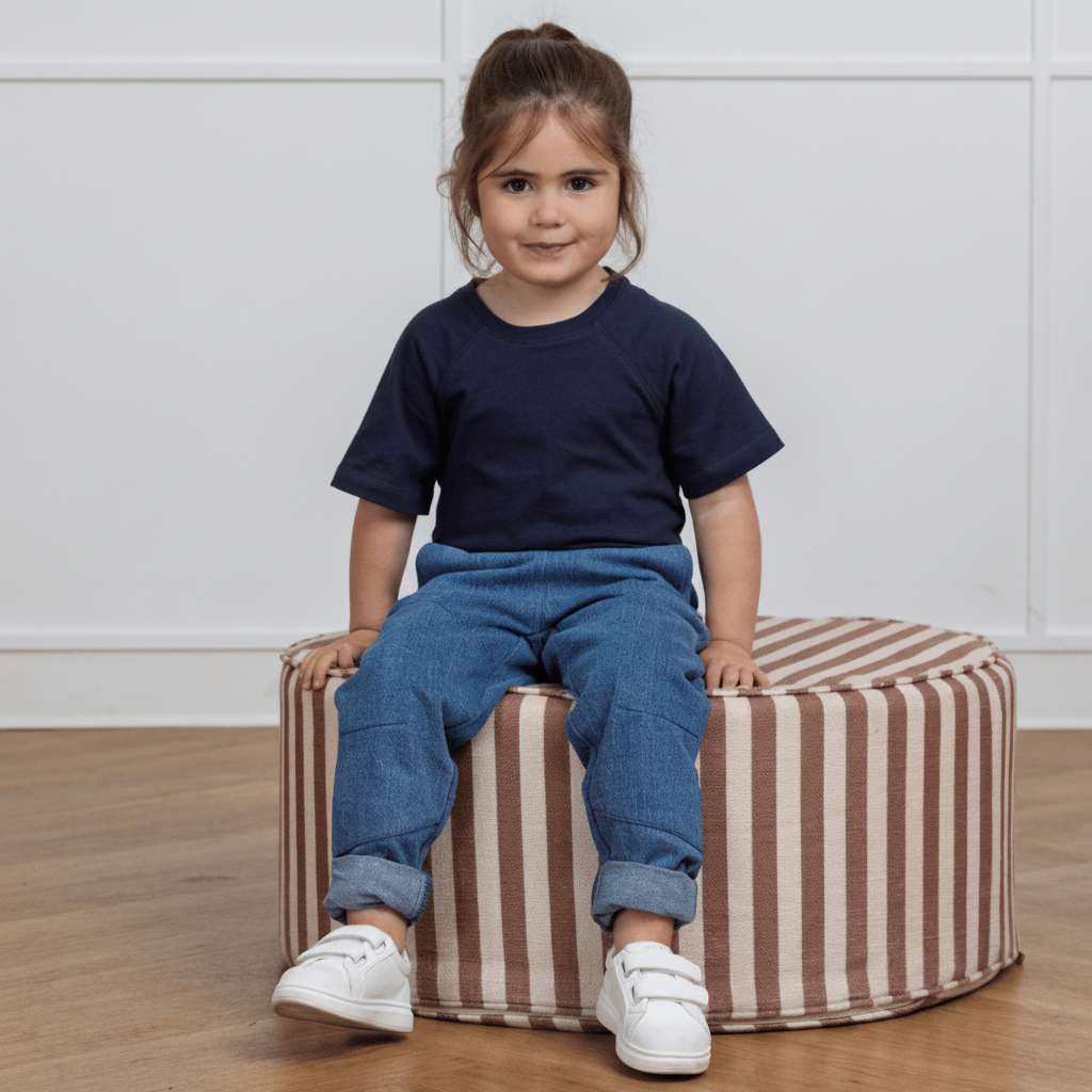 Toddler in navy tee and blue upcycled denim pants, sitting on striped ottoman, wearing white sneakers