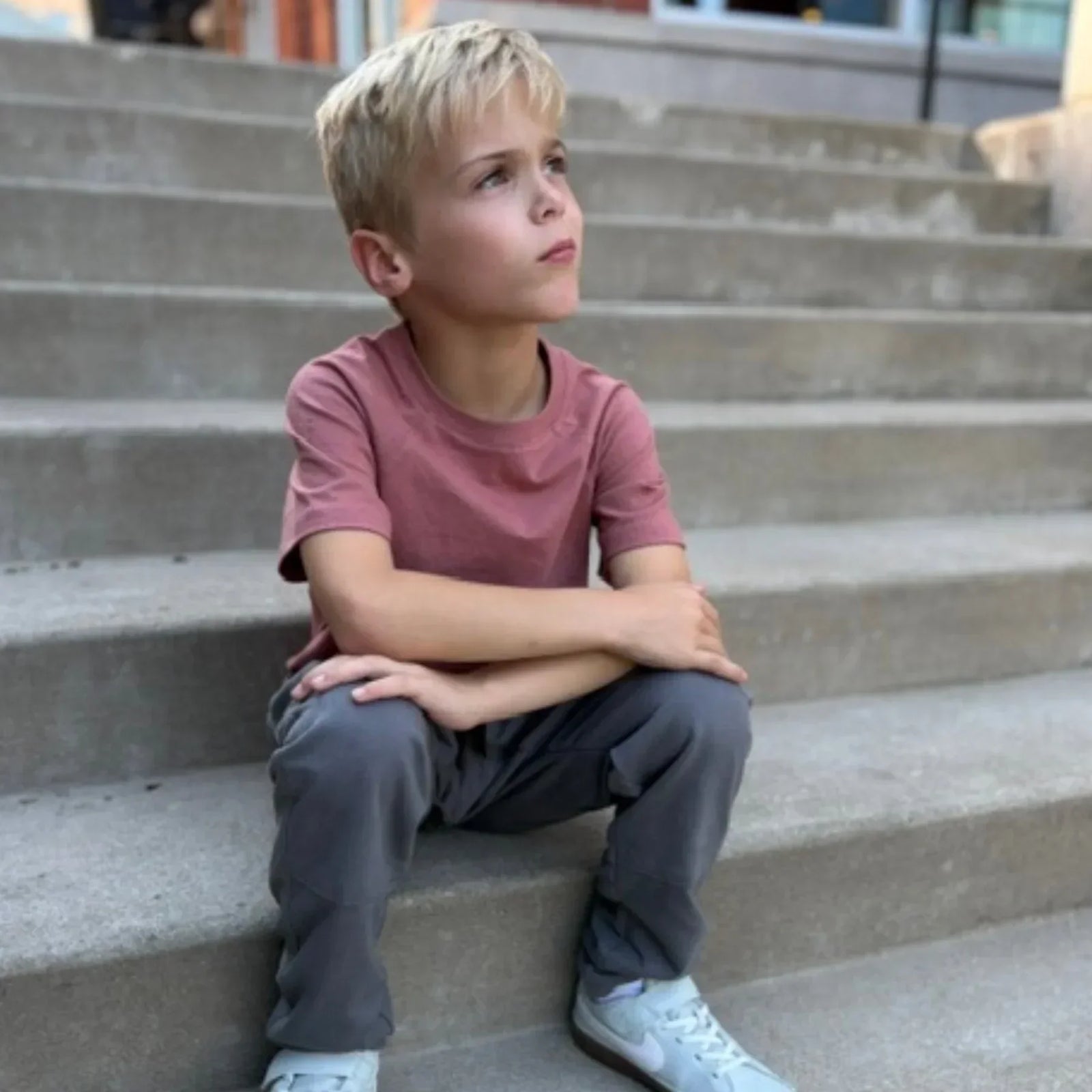 Blonde boy in pink t-shirt and gray pants by Jackalo sitting on outdoor steps