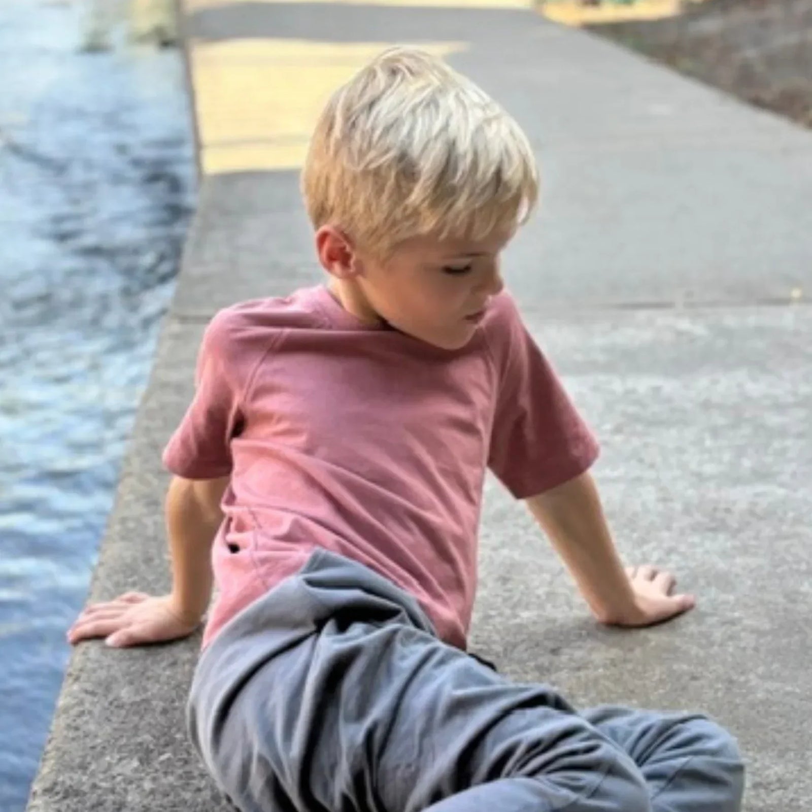 Young boy in pink t-shirt and gray pants from Jackalo sitting by water on a concrete path