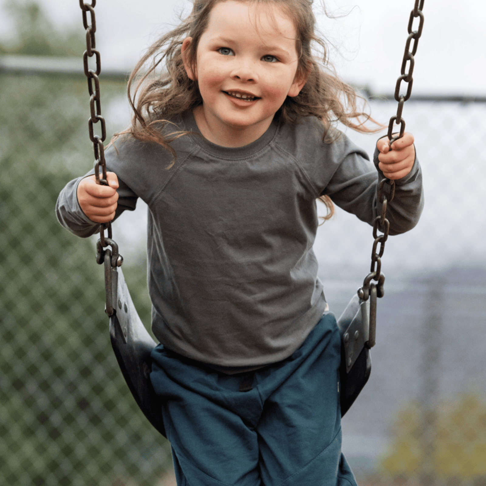 A young girl with long light brown hair smiling while riding a swing at an outdoor playground. She is wearing a dark gray long-sleeve raglan shirt and dark teal pants.