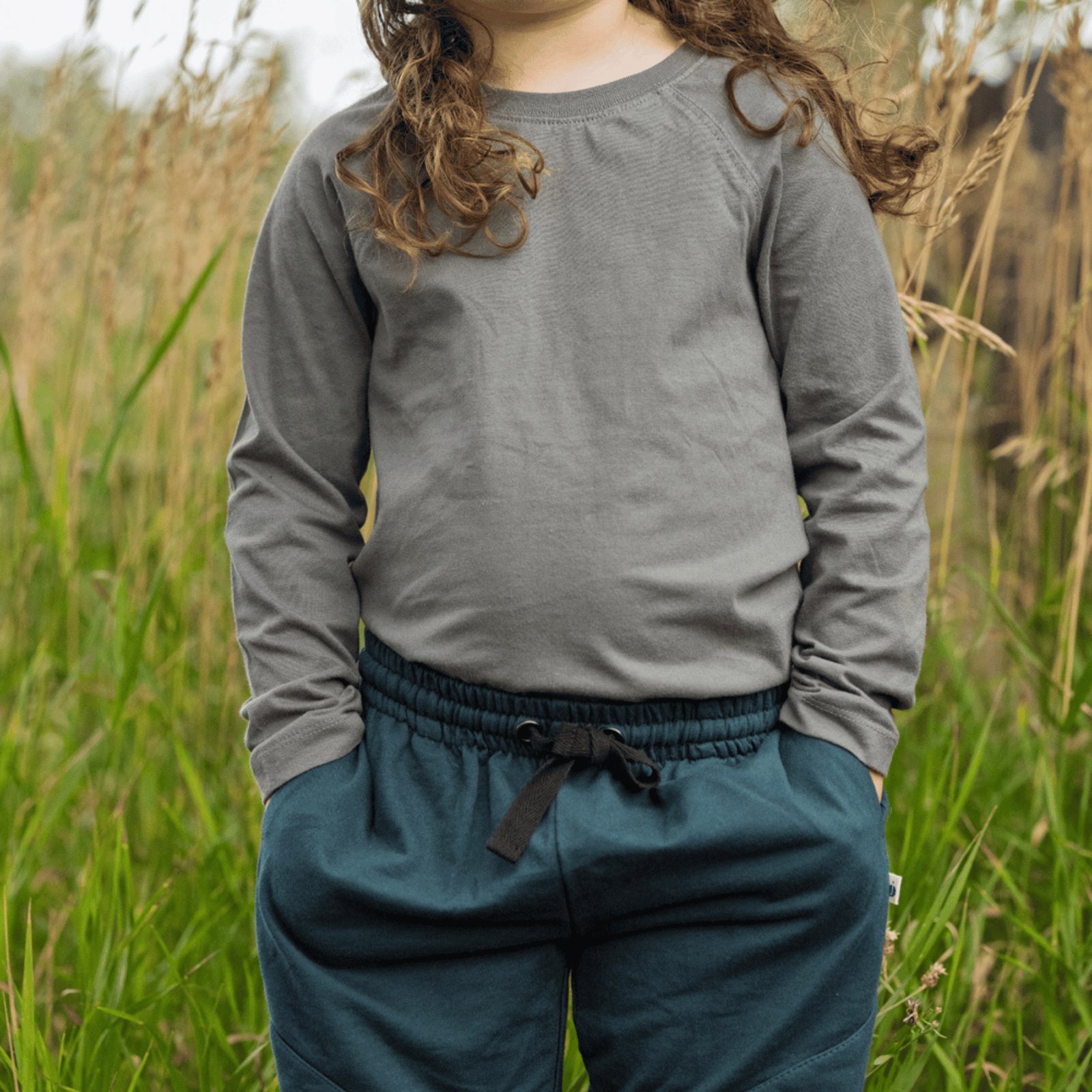 A child wearing a gray long-sleeve crewneck shirt and dark teal drawstring sweatpants, standing in a grassy outdoor field.