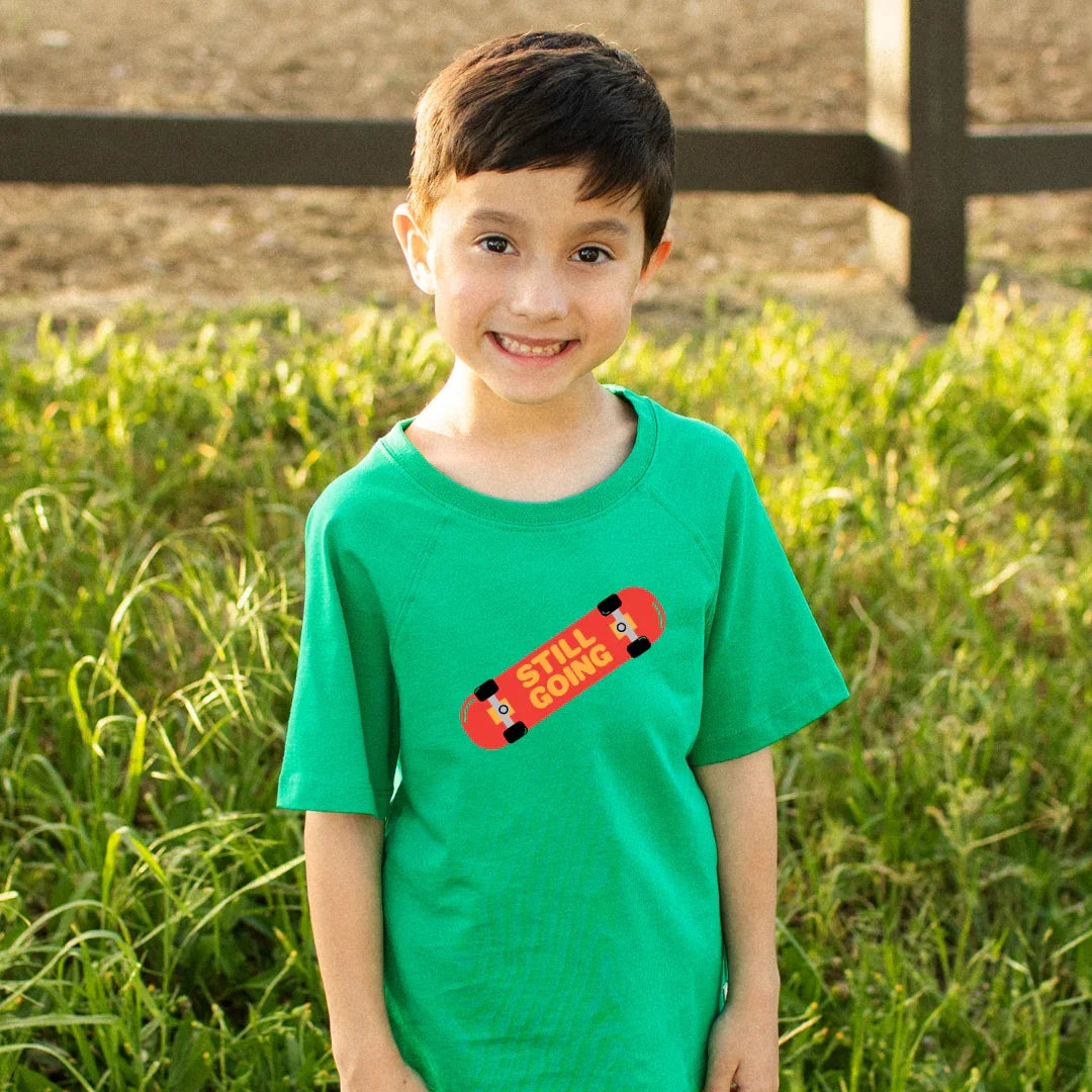 Boy in green tee with 'Still Going' skateboard graphic, outdoor grassy setting