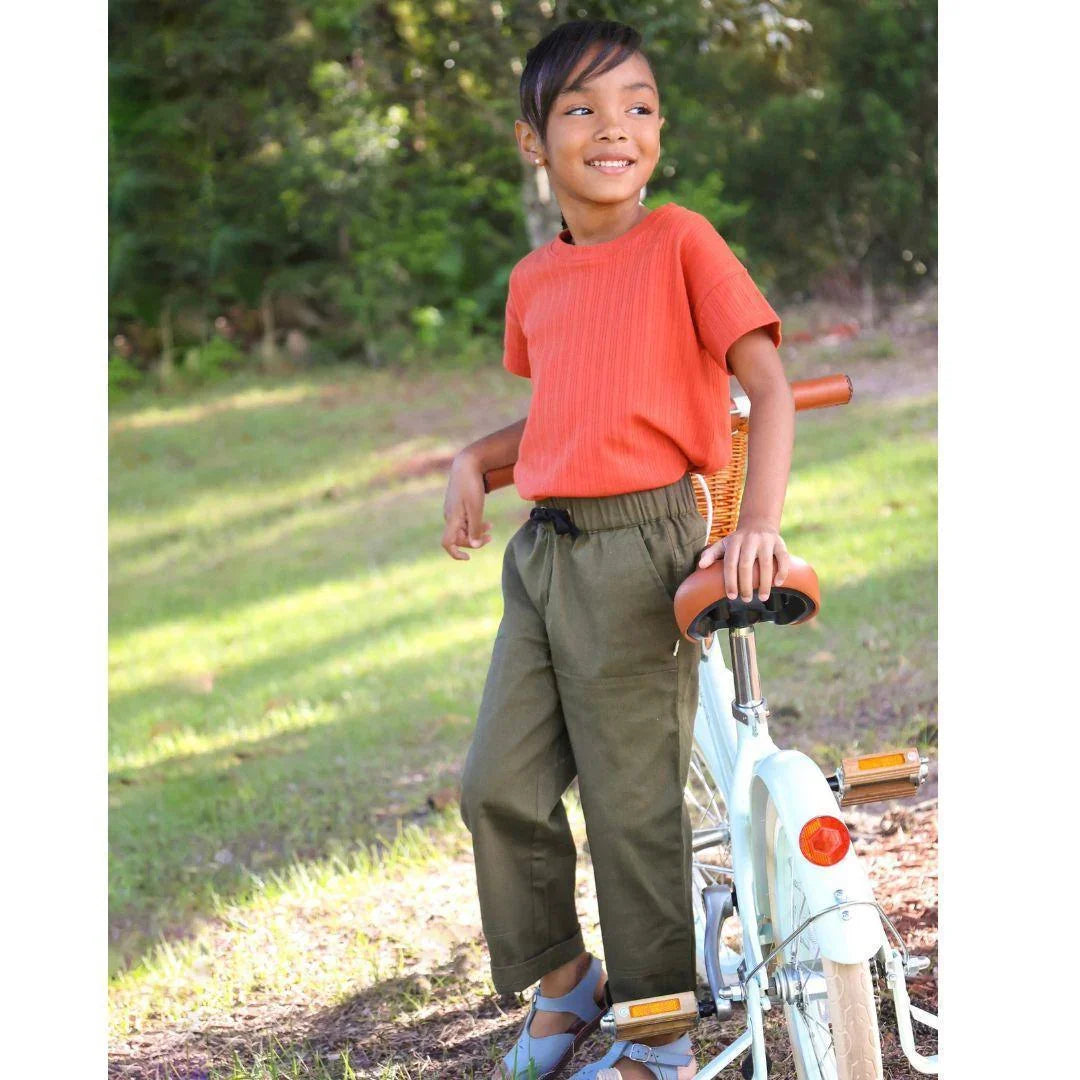 Child in sustainable dark olive pants by Jackalo, orange shirt, standing by bike outdoors