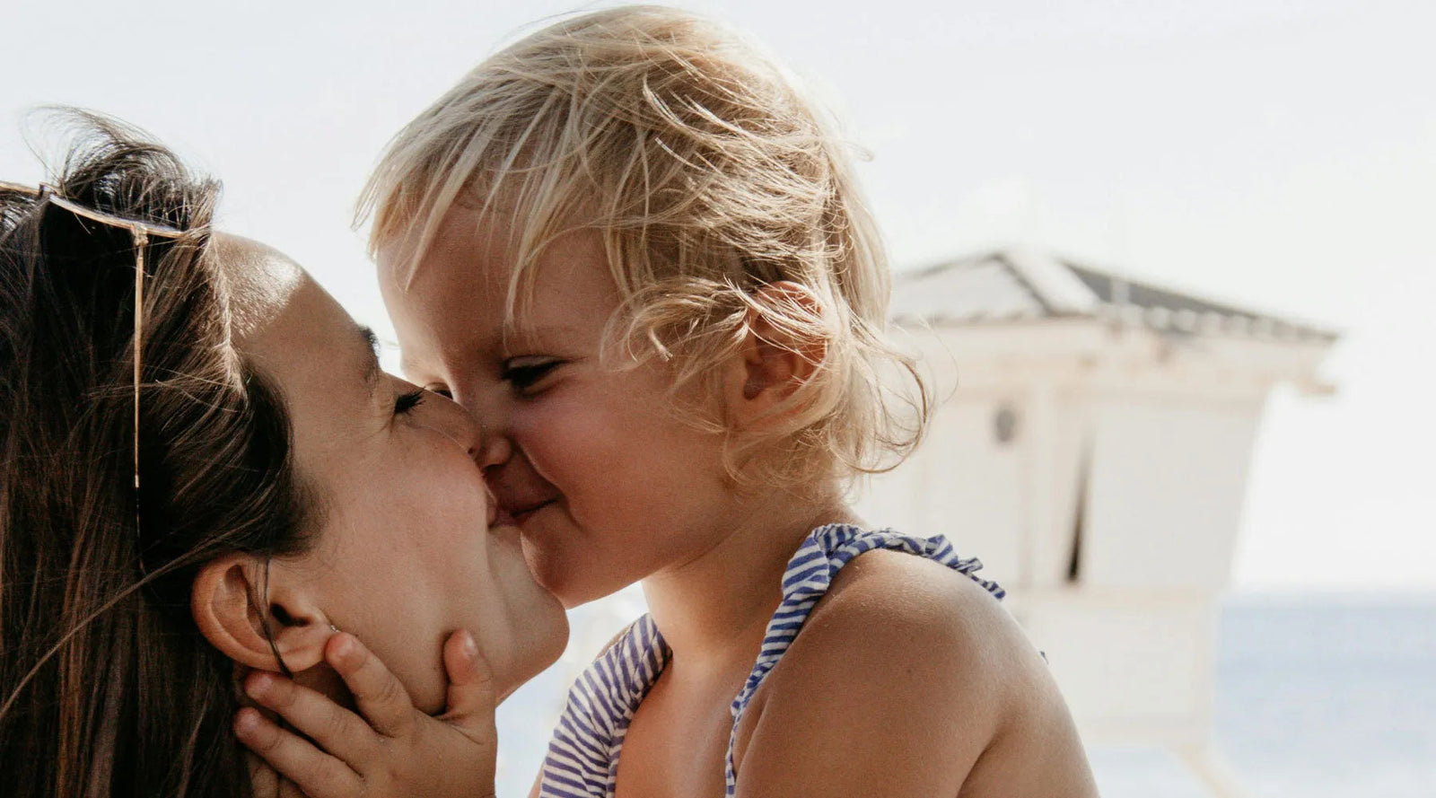 Smiling little girl in striped sundress kissing mother on a sunny beach near the sea, Jackalo.
