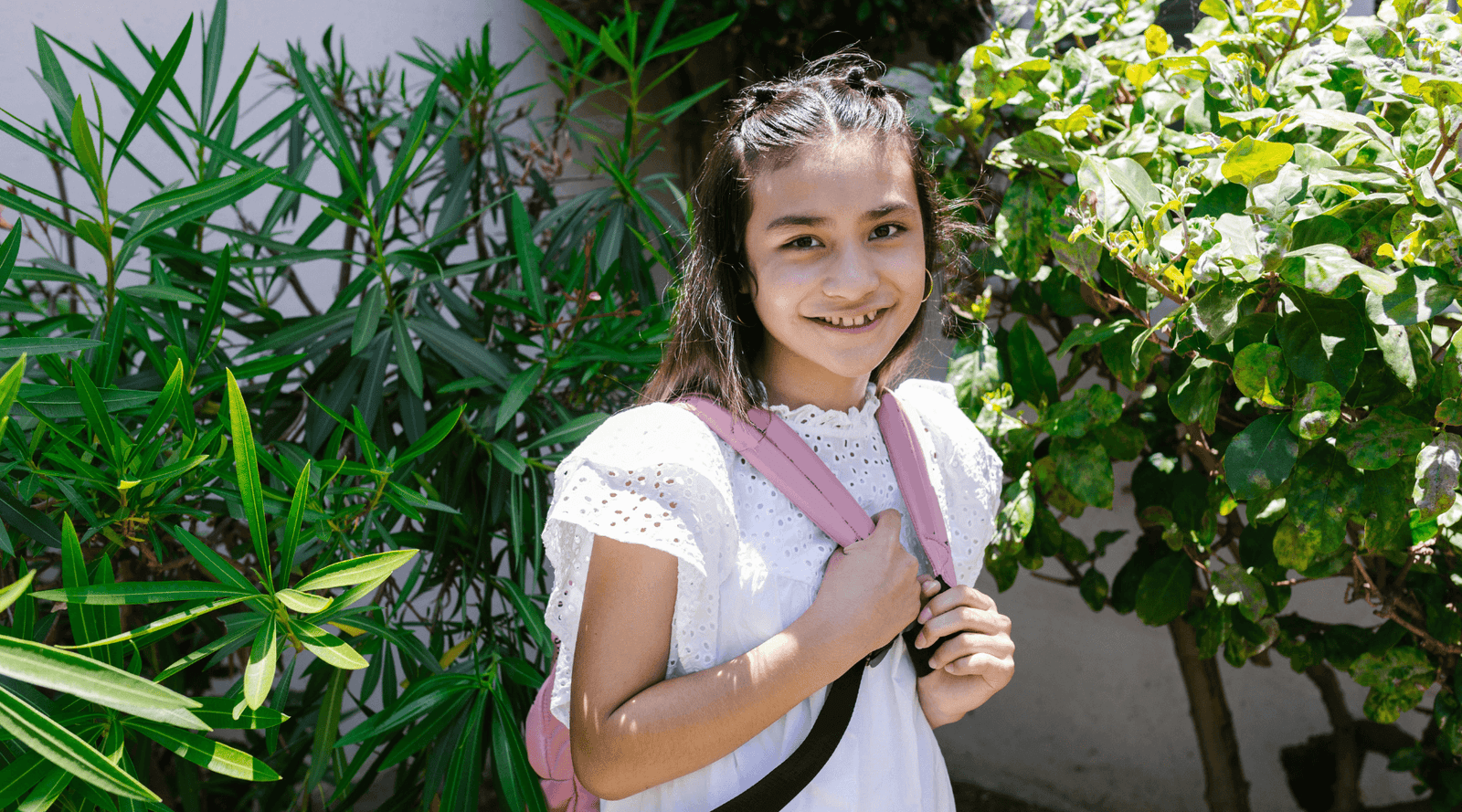 Smiling girl in white eyelet dress with pink backpack outdoors in green garden, sustainable kids clothing