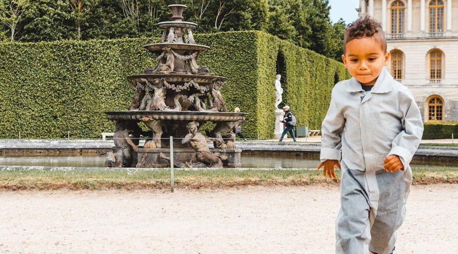 a young boy in coveralls runs in front of a fountain