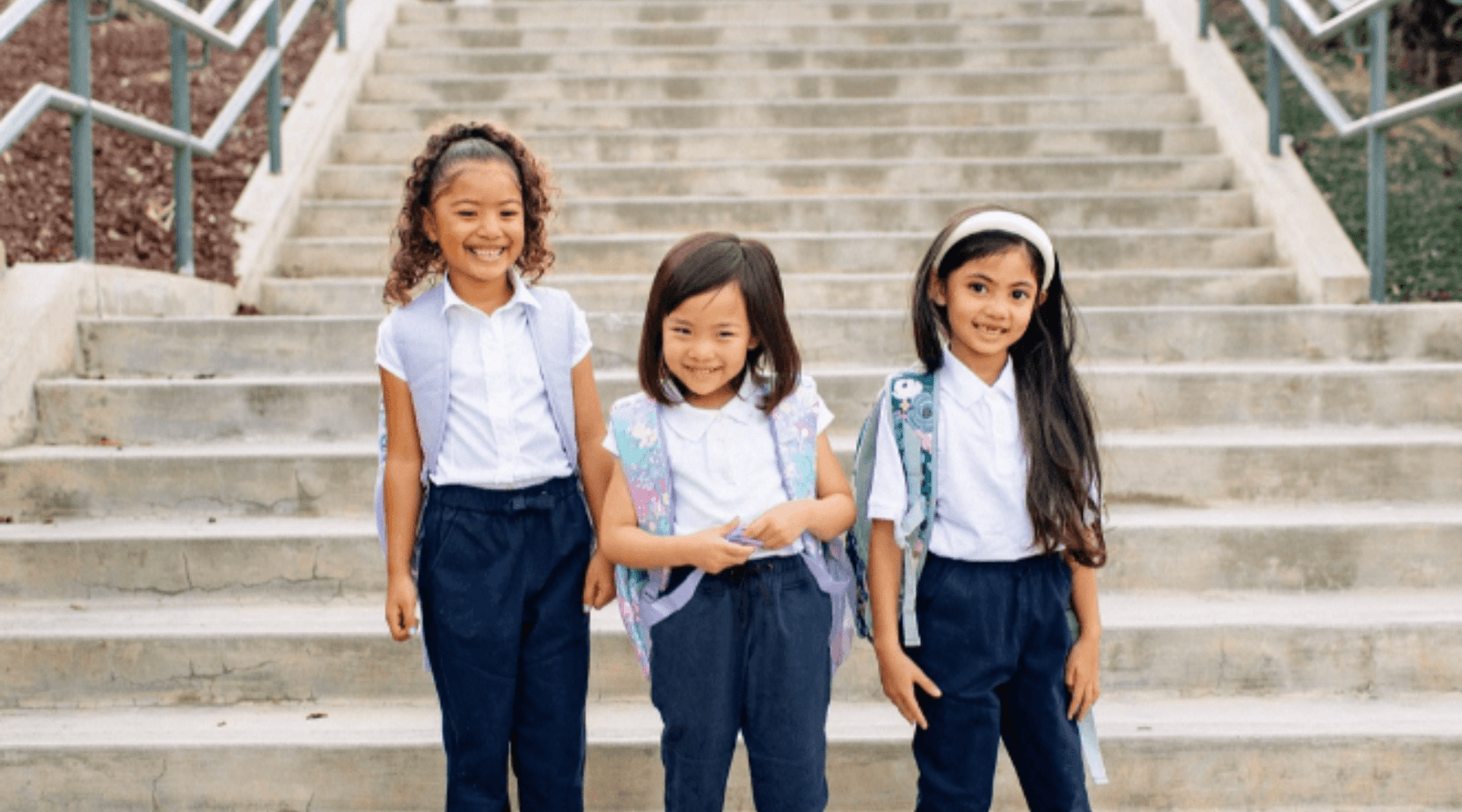 Three young girls in school uniforms and backpacks, smiling on outdoor steps, Jackalo kids clothes