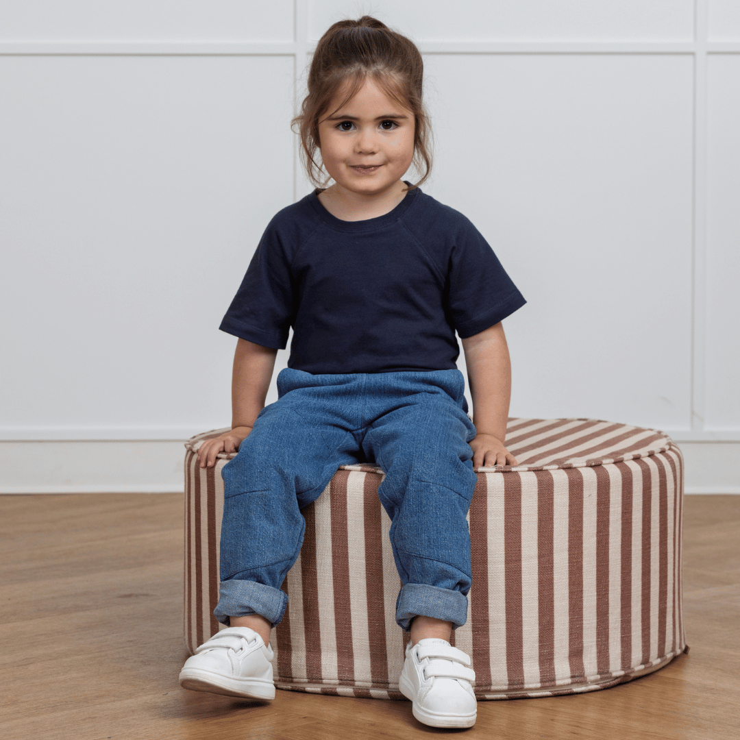Toddler in navy tee and blue denim pants, sitting on striped ottoman, wearing white sneakers