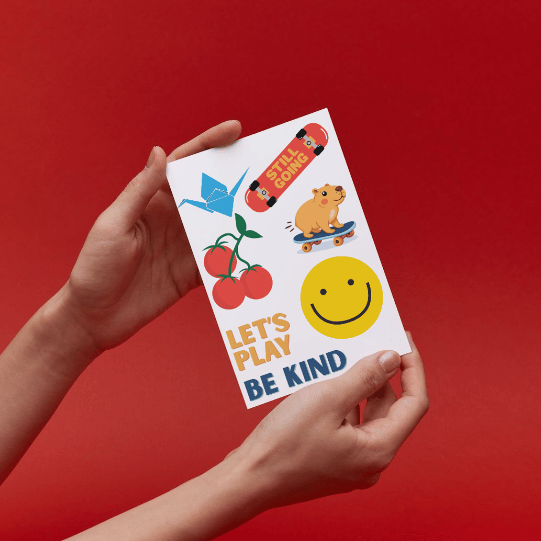 A person holds a stain cover sheet featuring colorful illustrations of a blue origami crane, red cherries, a yellow smiley face, a skateboard with the words 'LET'S PLAY', and a cartoon dog on a skateboard, with the text 'LET'S PLAY BE KIND'. The background is a solid bright red.