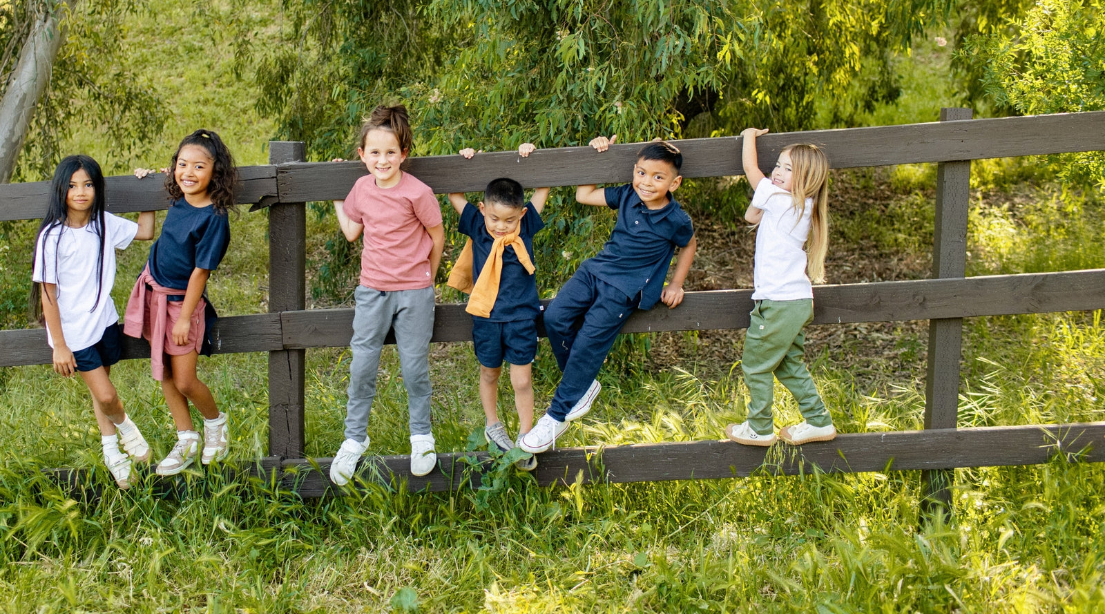 Children standing on a wooden fence in a grassy area with trees in the background