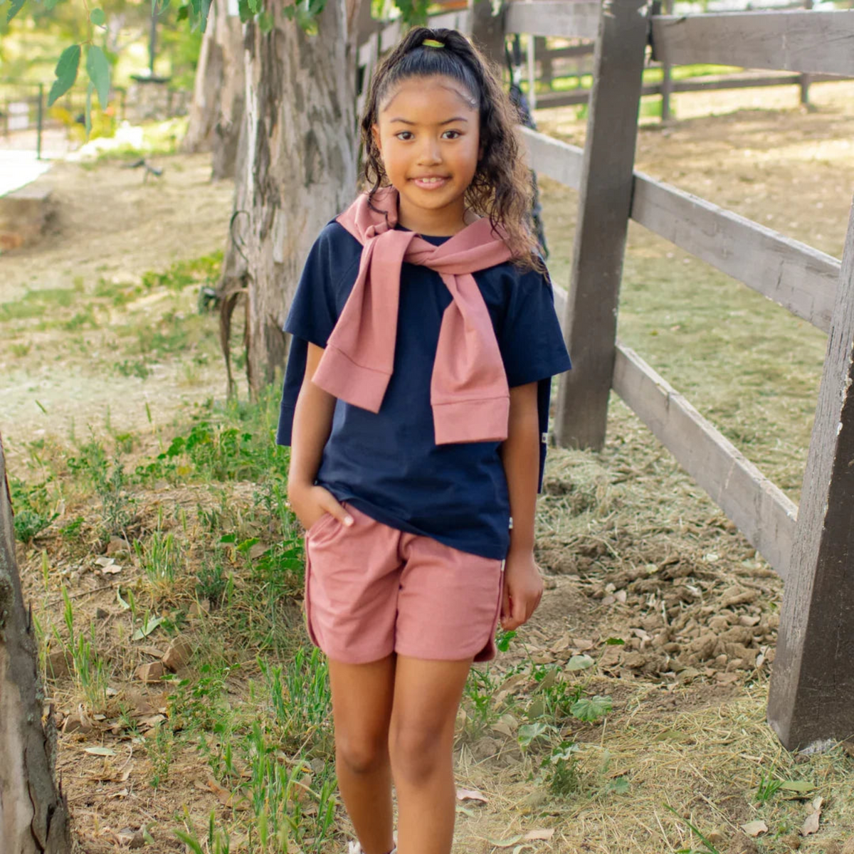 Girl in navy tee and pink shorts with matching scarf, standing outdoors near wooden fence