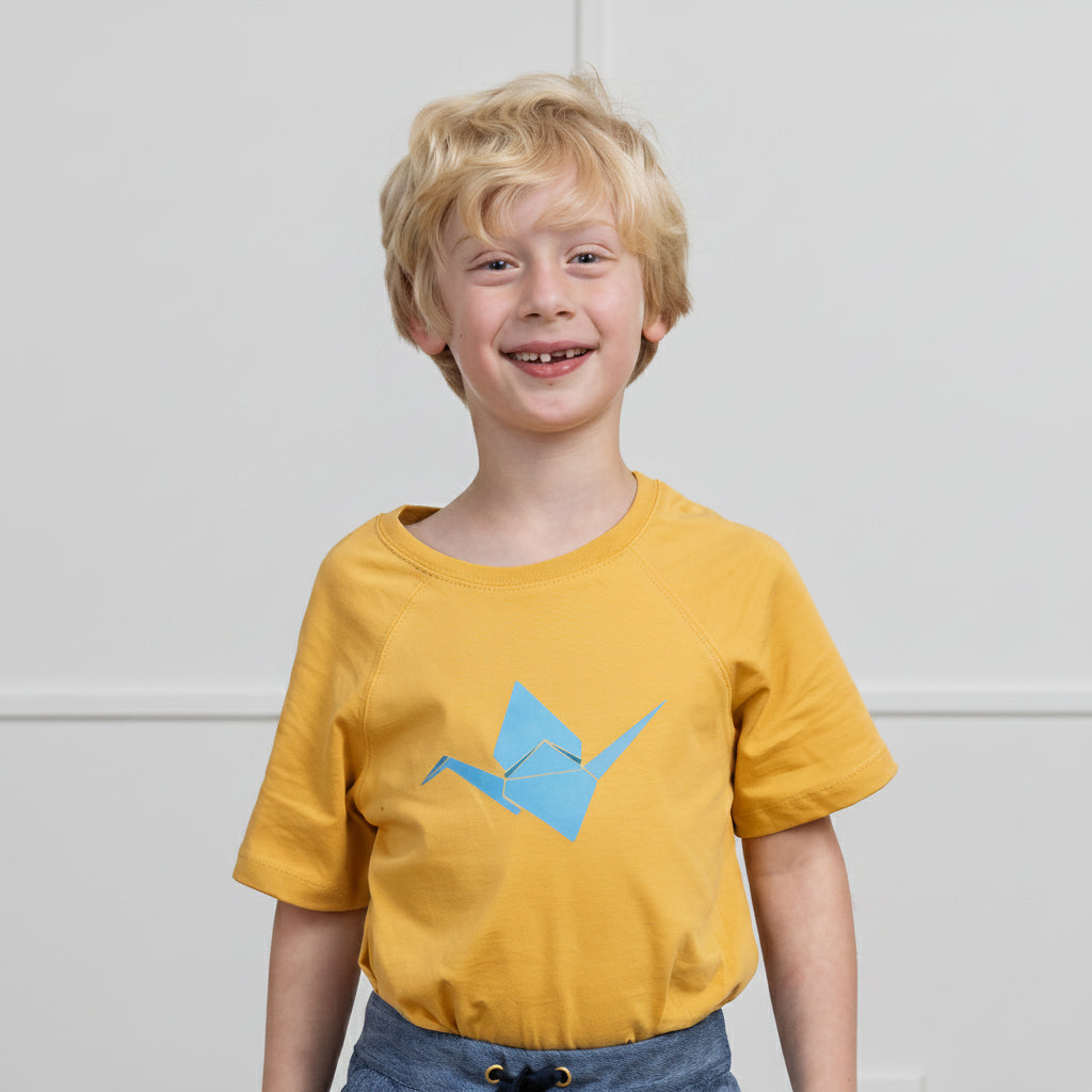 A young blonde boy smiling while wearing a mustard yellow t-shirt with a light blue origami crane graphic on the front, paired with blue jeans, set against a plain light gray wall.