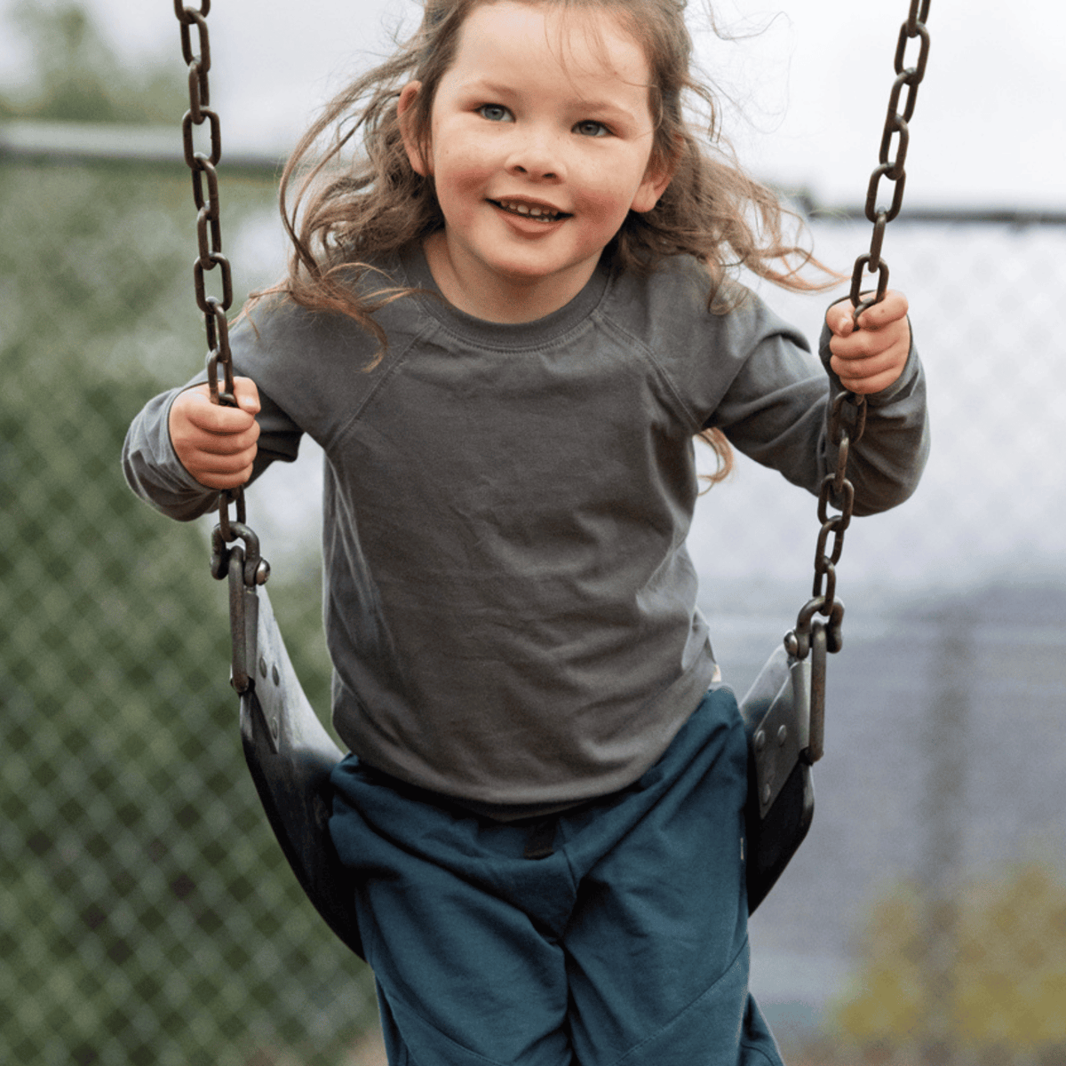 A young girl with long light brown hair smiling while riding a swing at an outdoor playground. She is wearing a dark gray long-sleeve raglan shirt and dark teal pants.