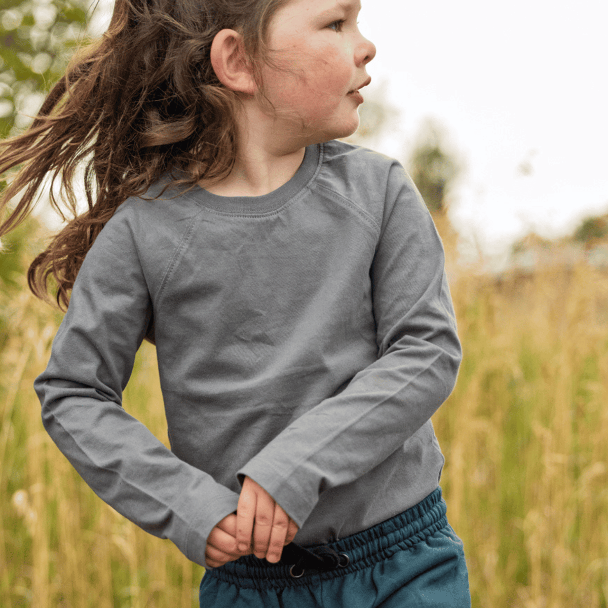 A young child wearing a gray long-sleeve crewneck shirt and teal pants, standing outdoors in a grassy field.