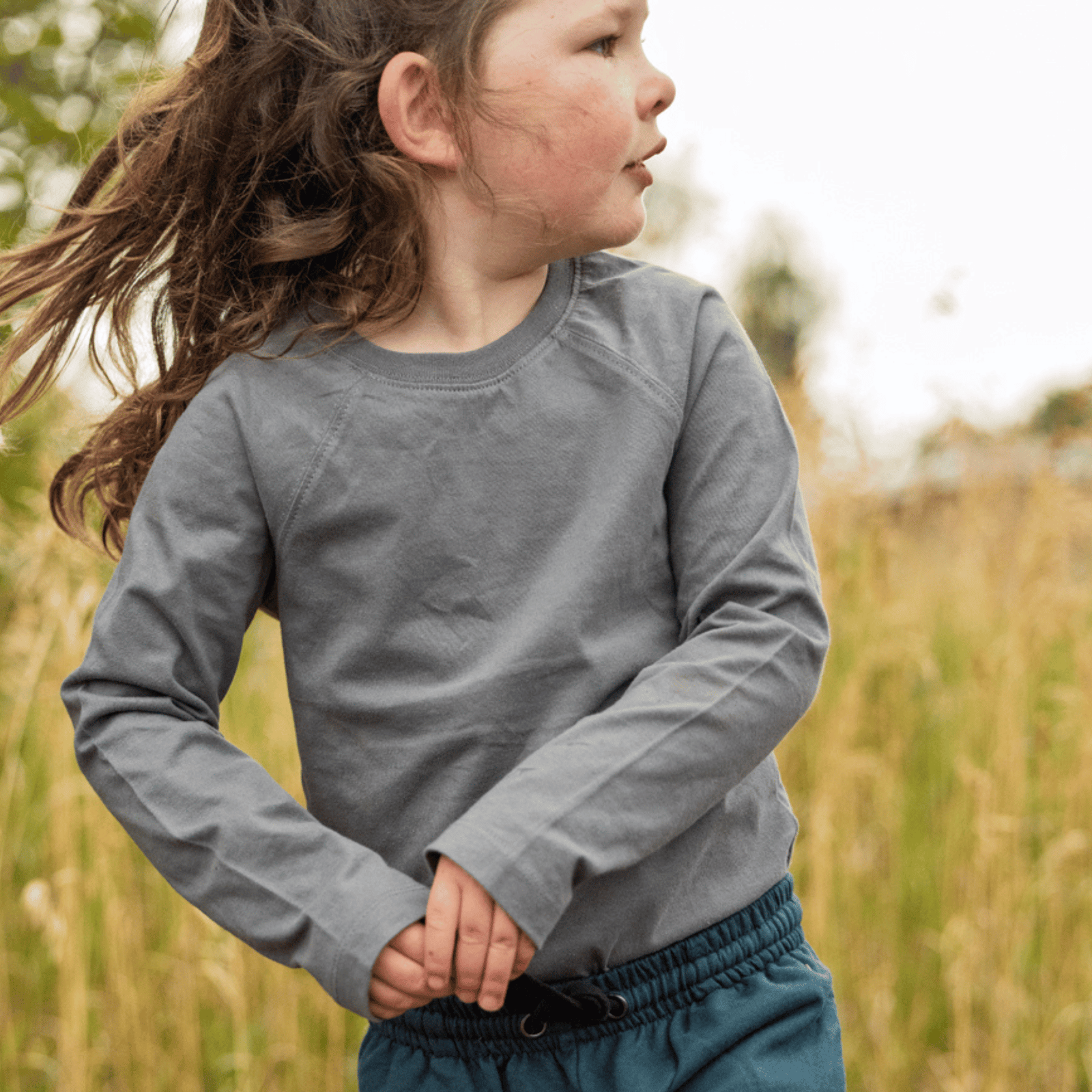 A young child wearing a gray long-sleeve crewneck shirt and teal pants, standing outdoors in a grassy field.