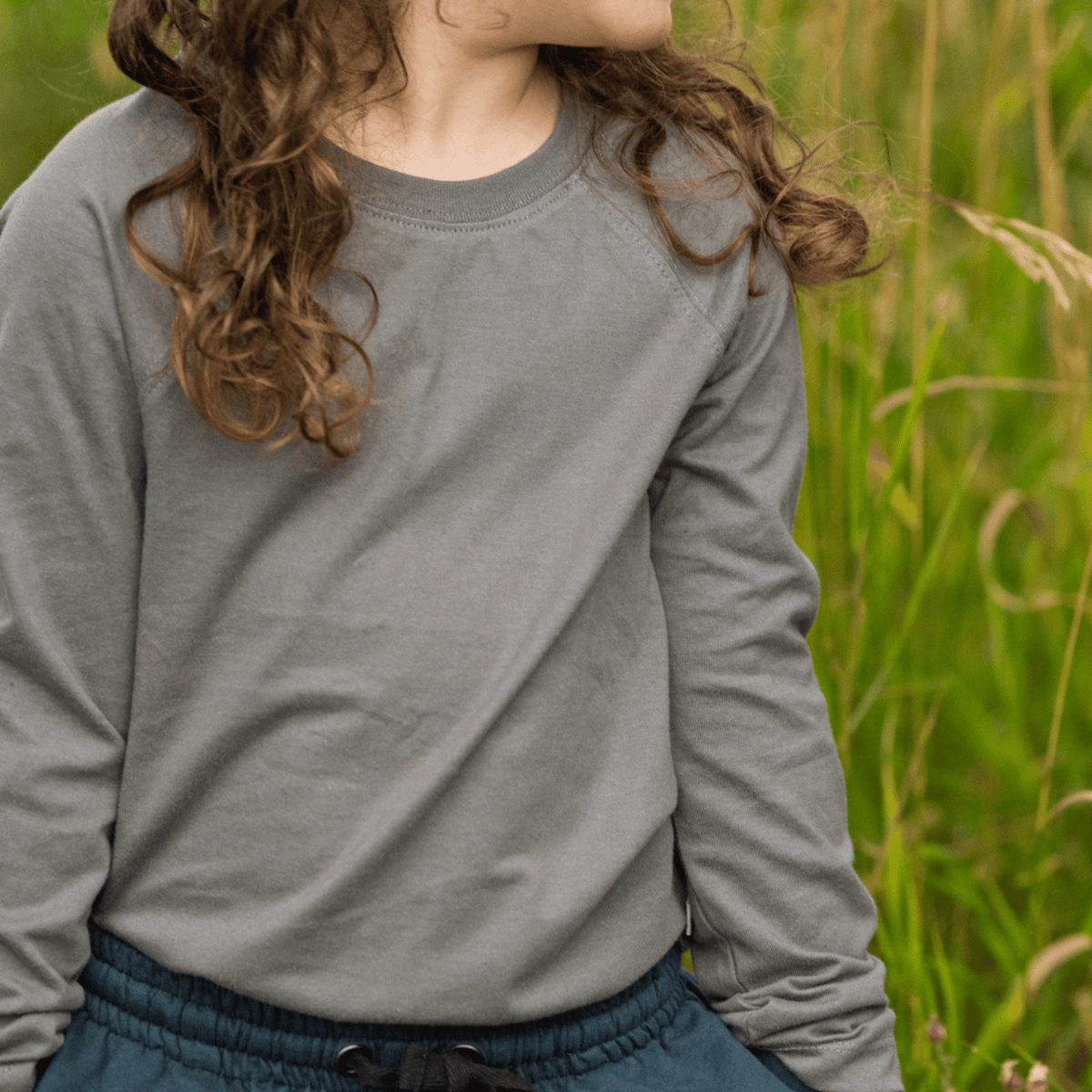 A child wearing a heather gray long-sleeve crewneck sweatshirt and dark teal drawstring pants, standing outdoors in a grassy field.