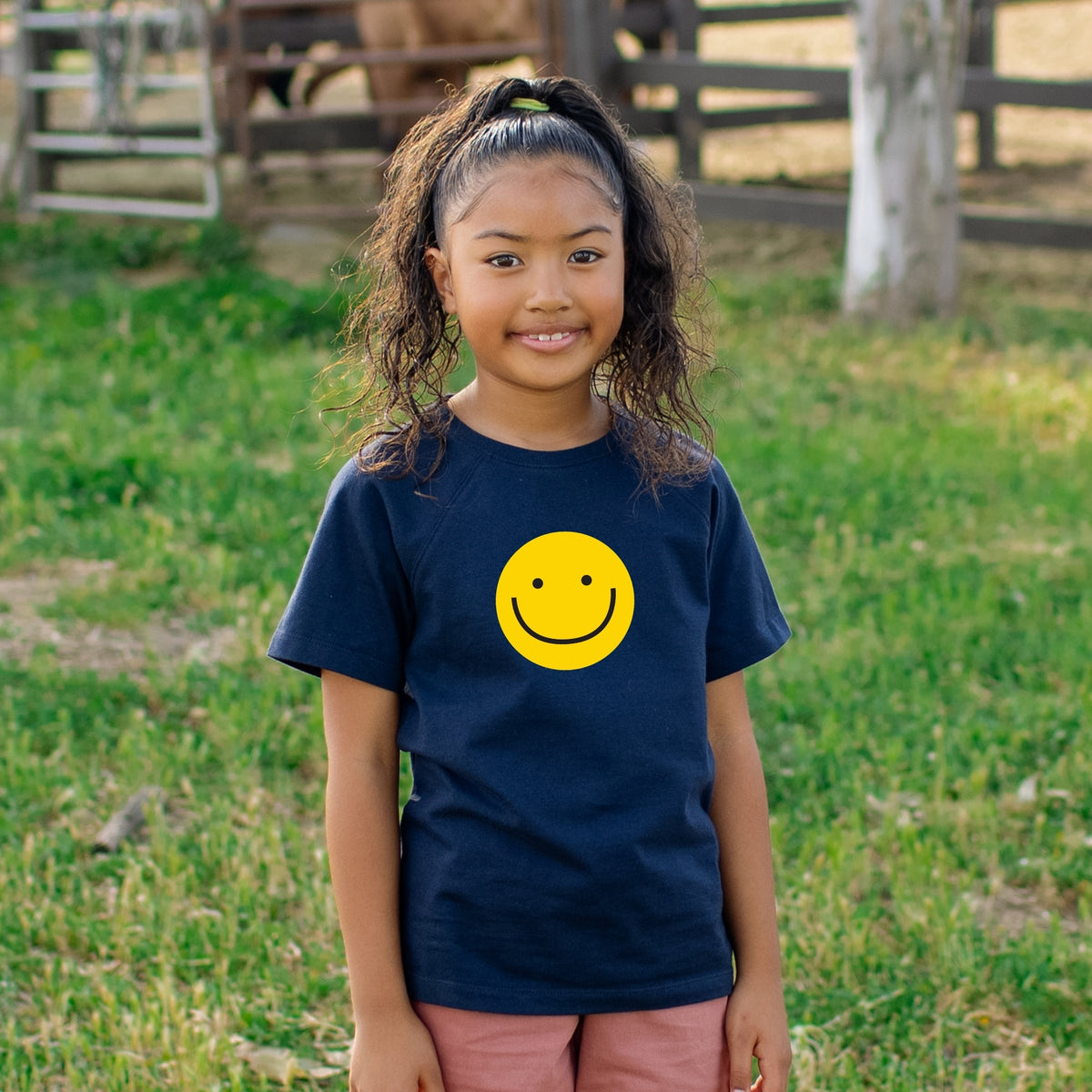 A young girl with curly hair stands outdoors on a grassy field wearing a navy blue t-shirt with a yellow smiley face graphic on the chest.