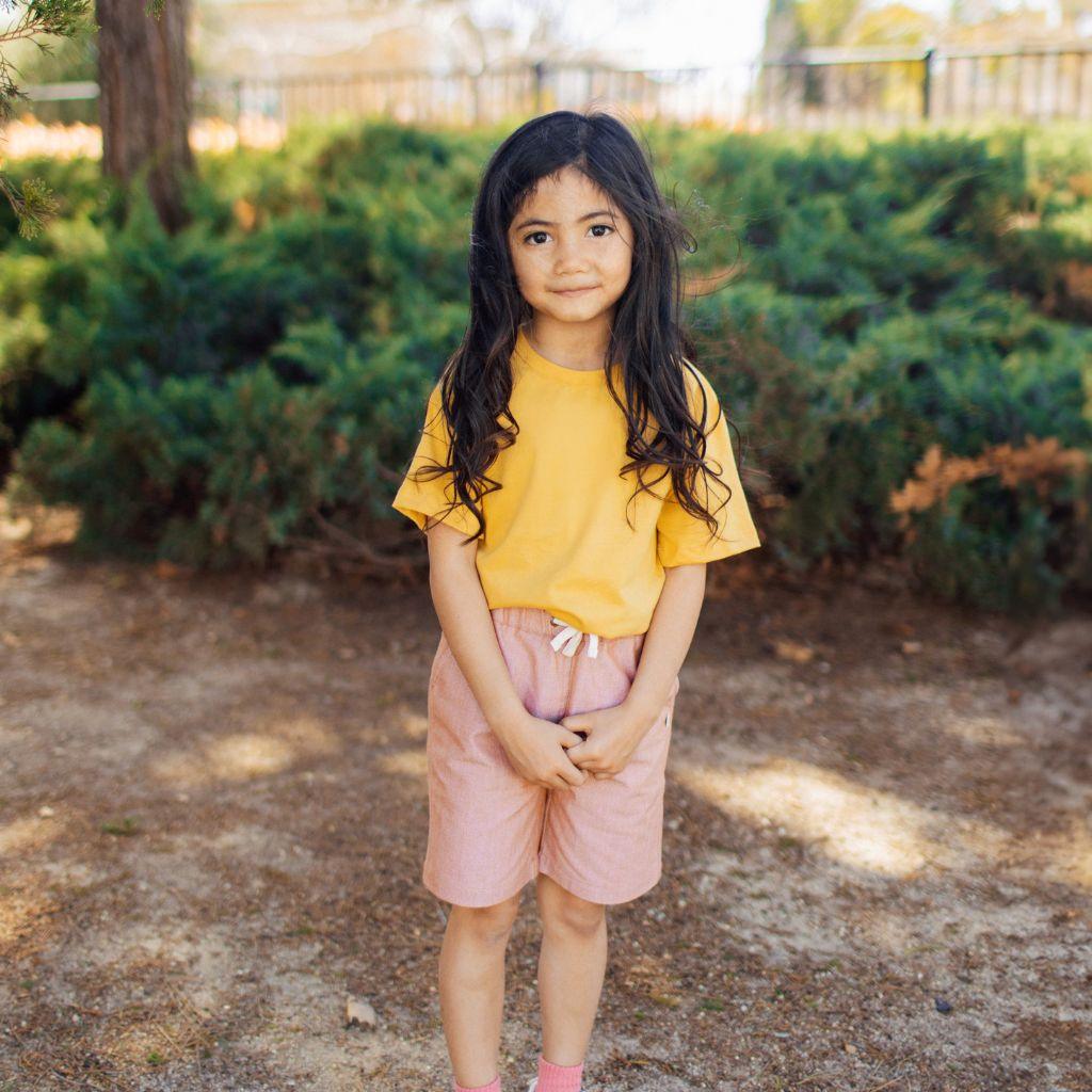 A girl standing wearing a yellow shirt and peach shorts