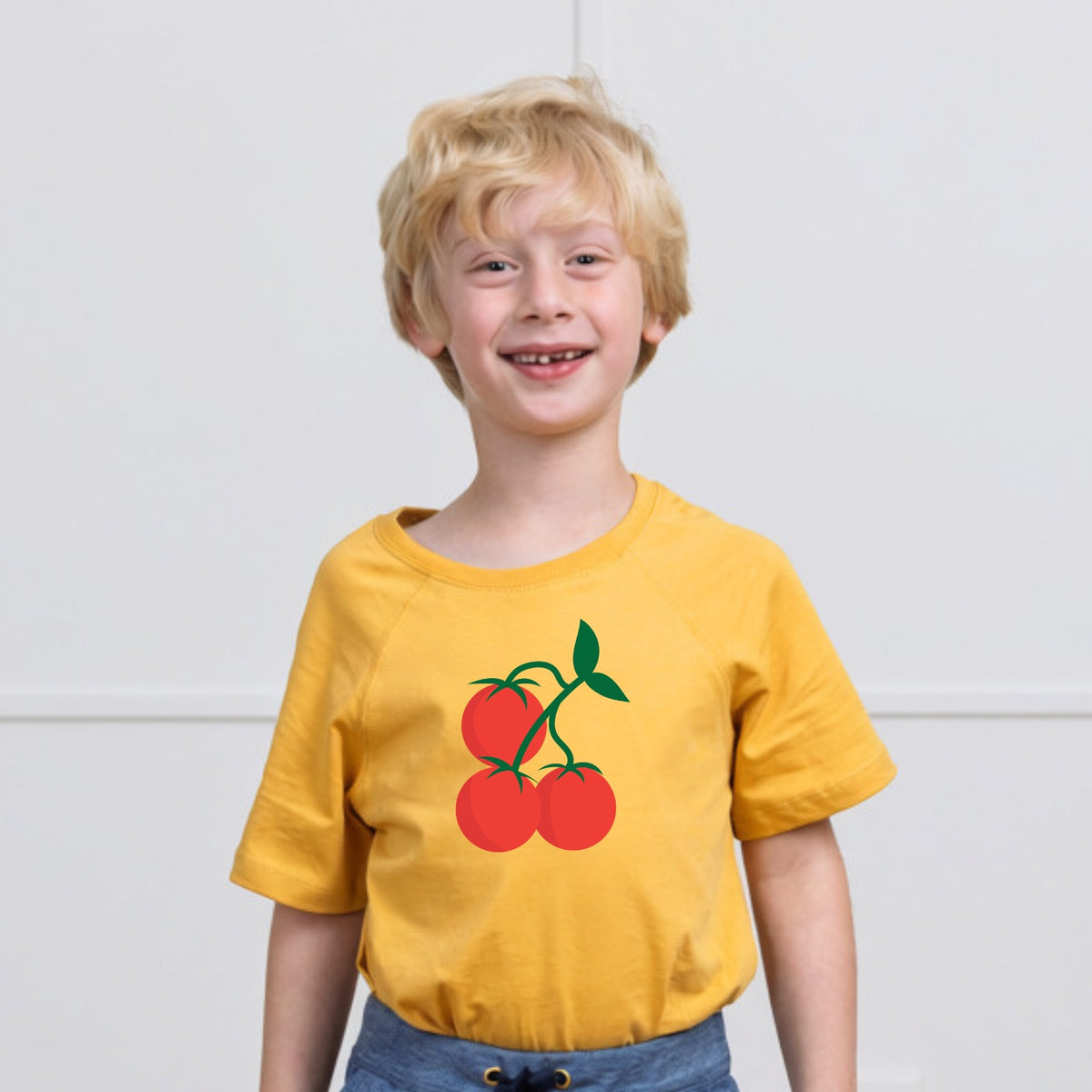 A young blonde boy smiling while wearing a yellow organic short-sleeve t-shirt with a graphic of three red tomatoes on the chest, paired with blue jeans, set against a plain white wall.