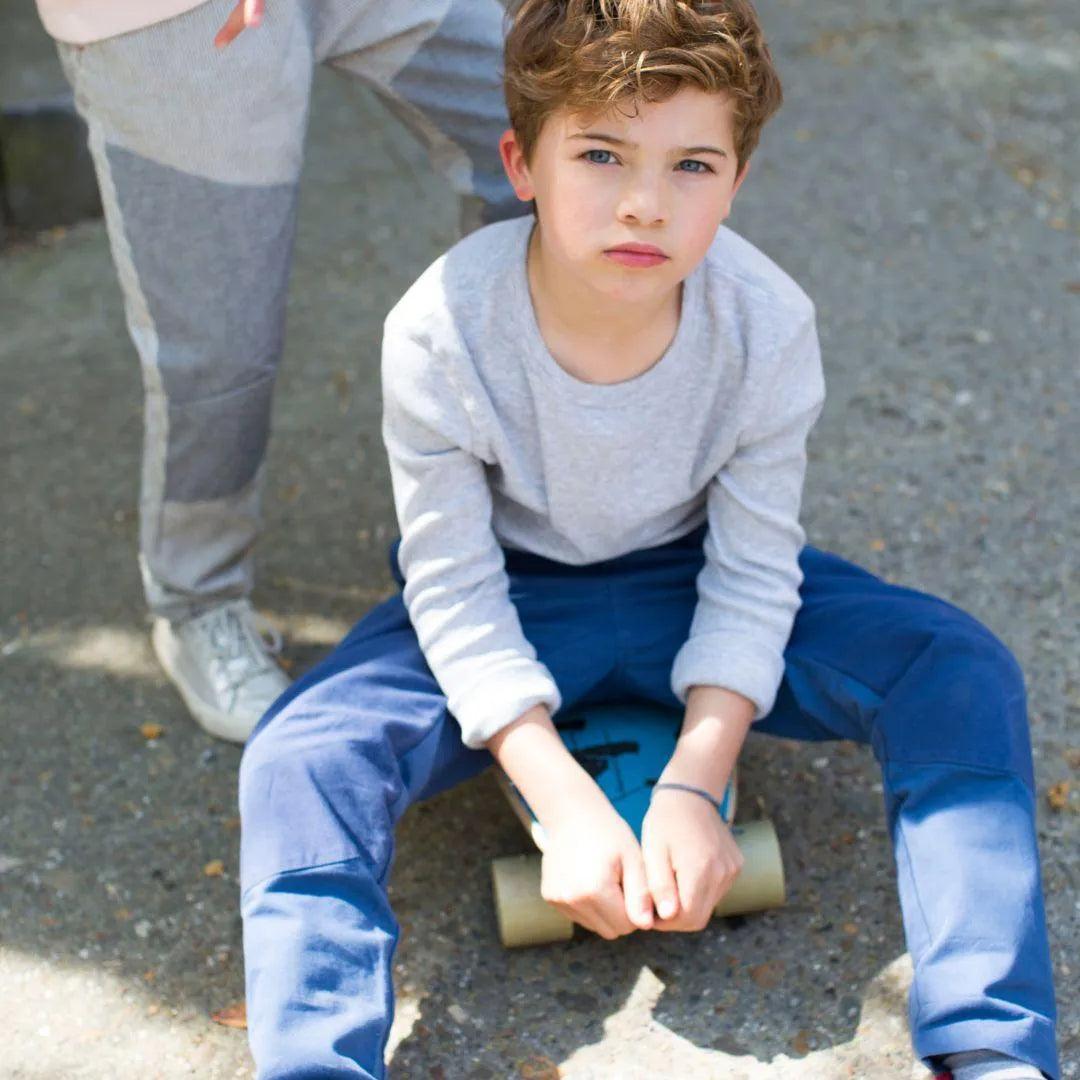 A young boy wearing blue Ash Pants sitting on a skateboard
